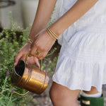 Person in white dress and green boots holding a brass watering can in a garden setting