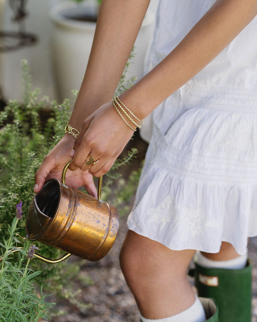 Person in white dress and green boots holding a brass watering can in a garden setting